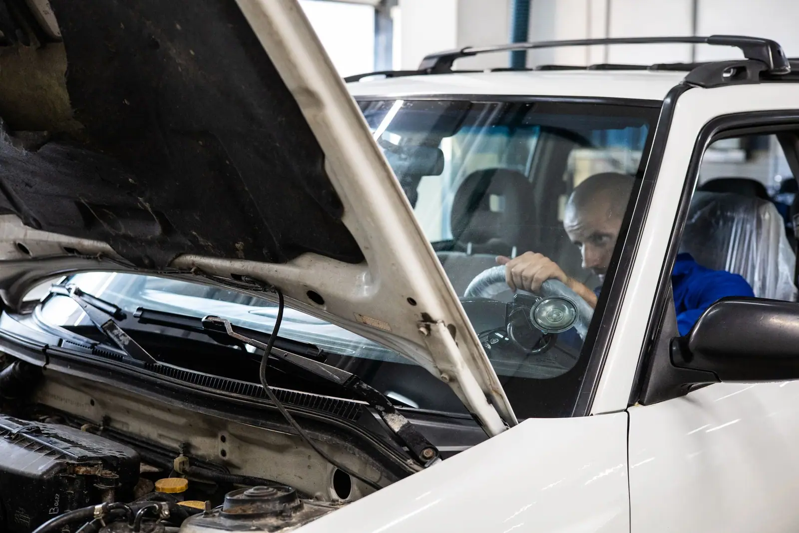 A mechanic in blue clothing looking into a car with its hood raised
