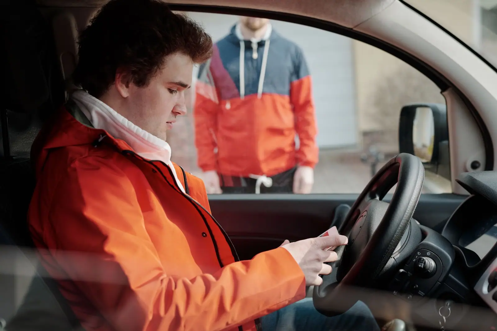 A woman in orange work coveralls seated in a car while another person stands outside