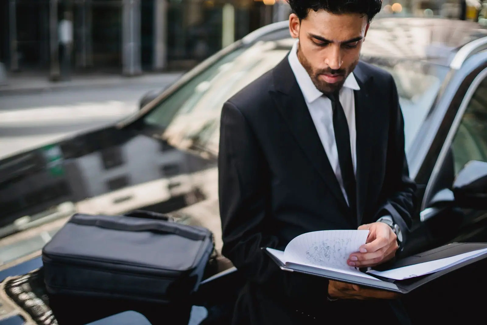 A man in a dark suit reviewing paperwork beside parked cars
